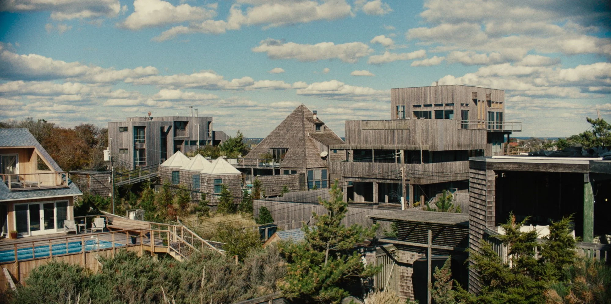Multiple modern and traditional houses with wooden exteriors, some with balconies and a swimming pool, set among trees under a partly cloudy sky.