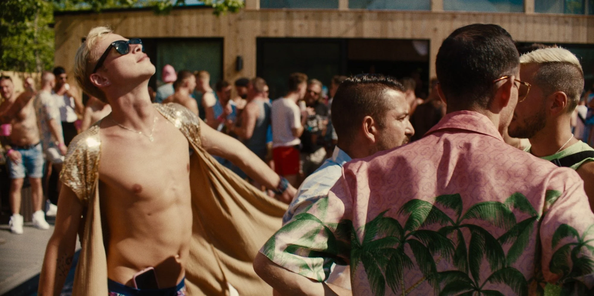 A shirtless person wearing sunglasses and a gold jacket, looking up with arms outstretched, in a crowded outdoor event with many people in the background.