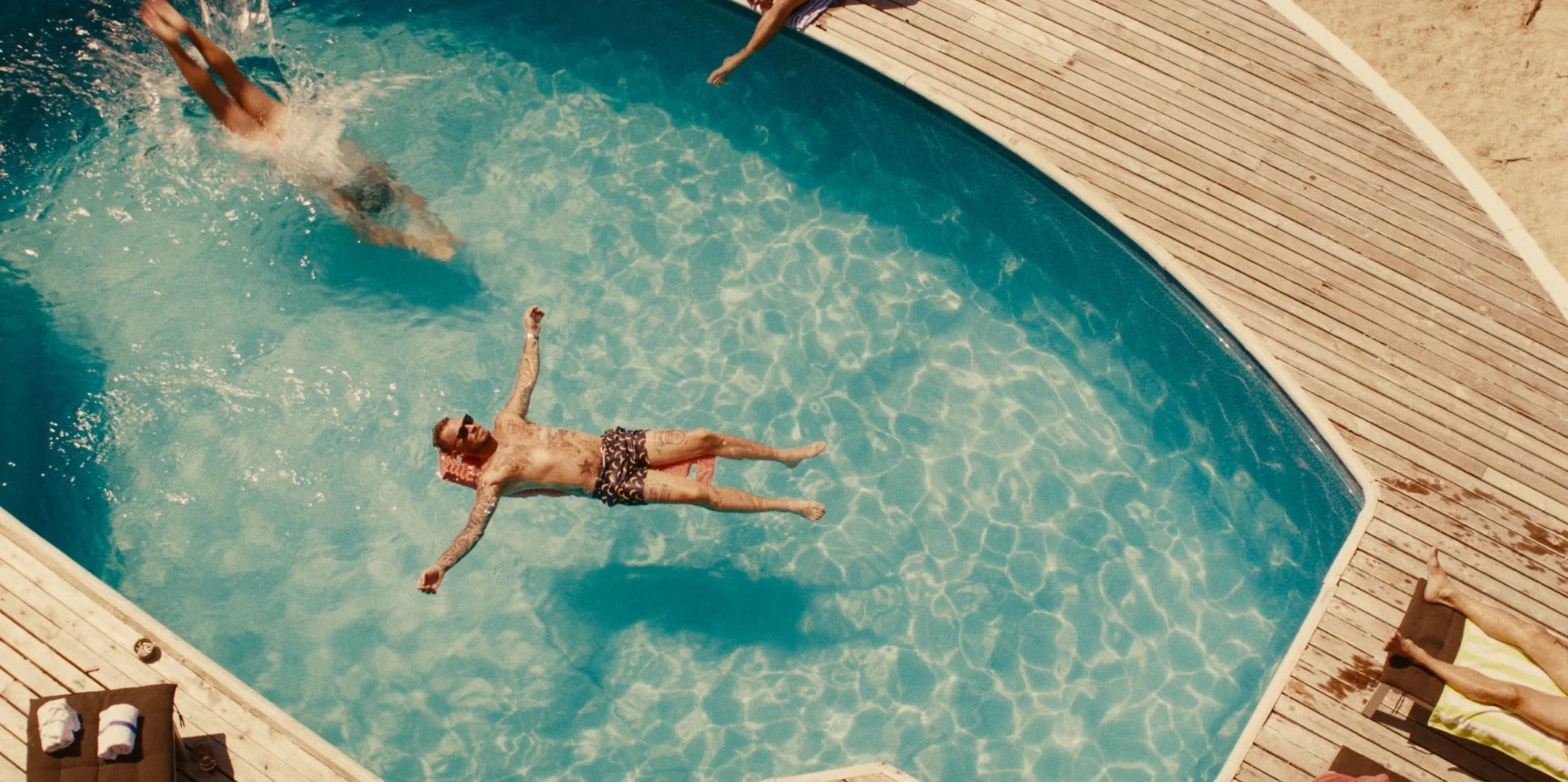 A man floating on his back in a swimming pool, wearing sunglasses and swim shorts, with a smile on his face. There are some towels and a lounge chair on the pool deck nearby.
