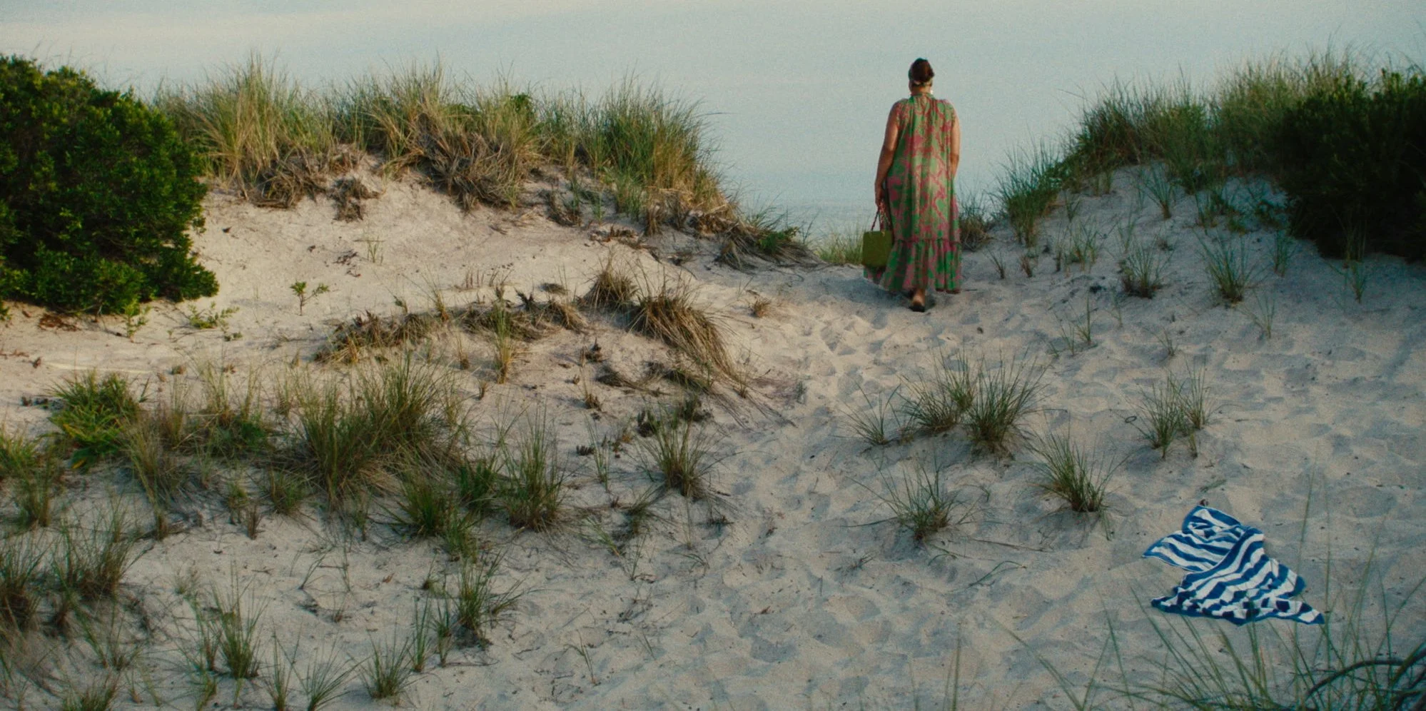 A woman in a long colorful dress walks on sandy dunes towards the water, carrying a green bag, with a blue and white striped towel on the sand nearby.