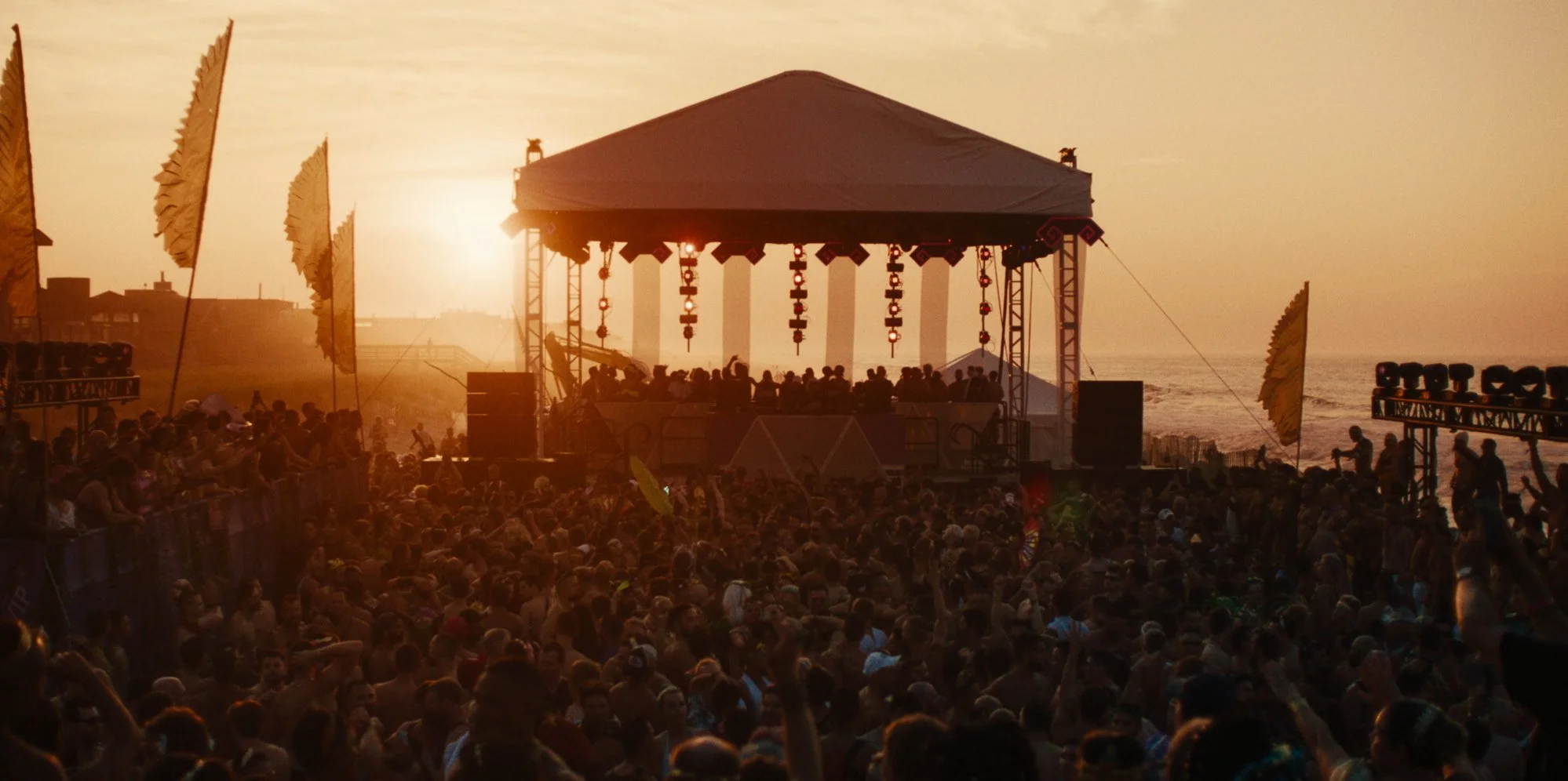 A large crowd gathered in front of a stage on the beach during sunset, with the ocean visible in the background. The stage is set up with lighting and decorations, and people are dancing and enjoying a live music event.