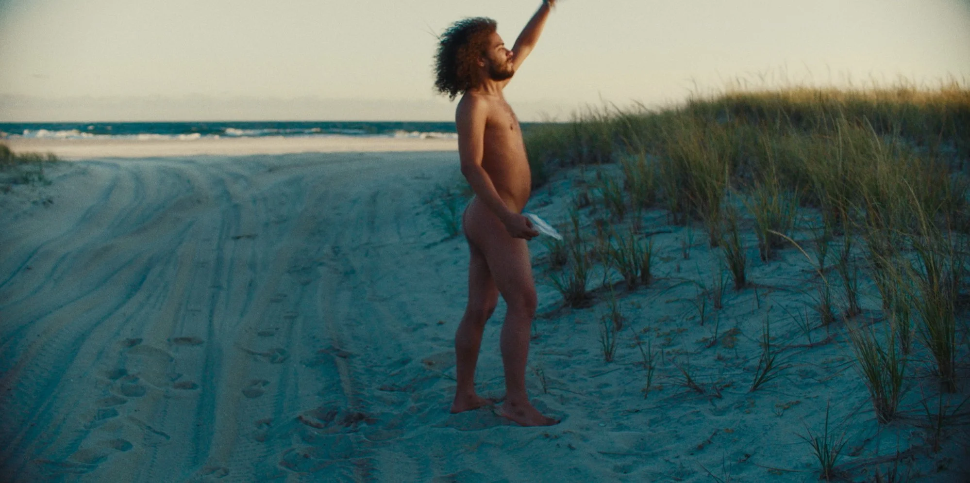 A man with curly hair and a beard standing on a sandy beach near grass, raising one arm, holding a small object in his other hand, with ocean waves in the background during sunset.