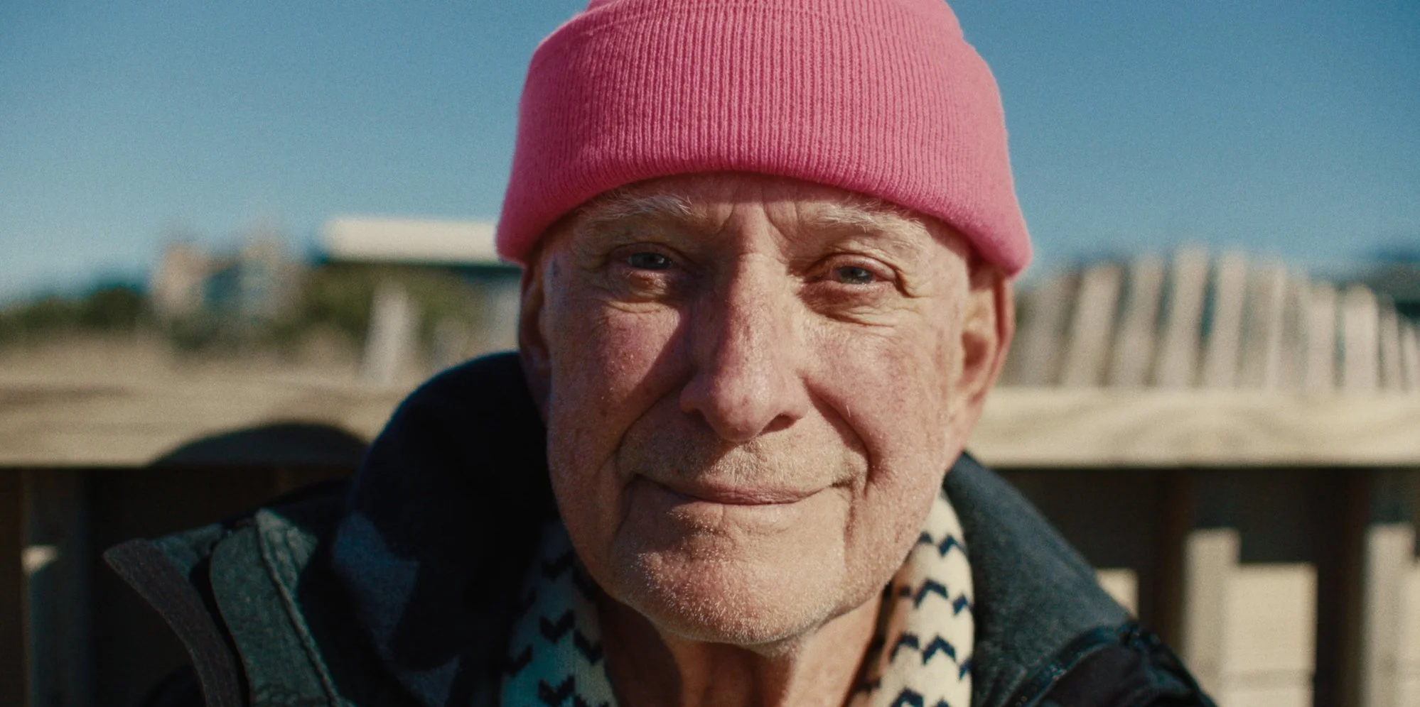 Close-up of an elderly man smiling outdoors, wearing a pink beanie and a dark jacket, with a blurry background of a boardwalk or beach.