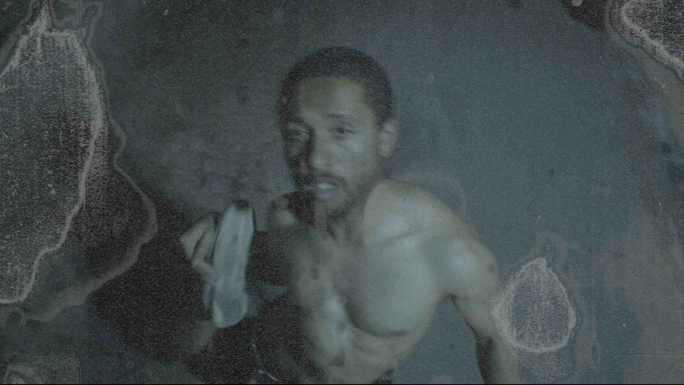 A young man with short curly hair, shirtless, holding a towel, standing behind a foggy glass surface nearest the camera.