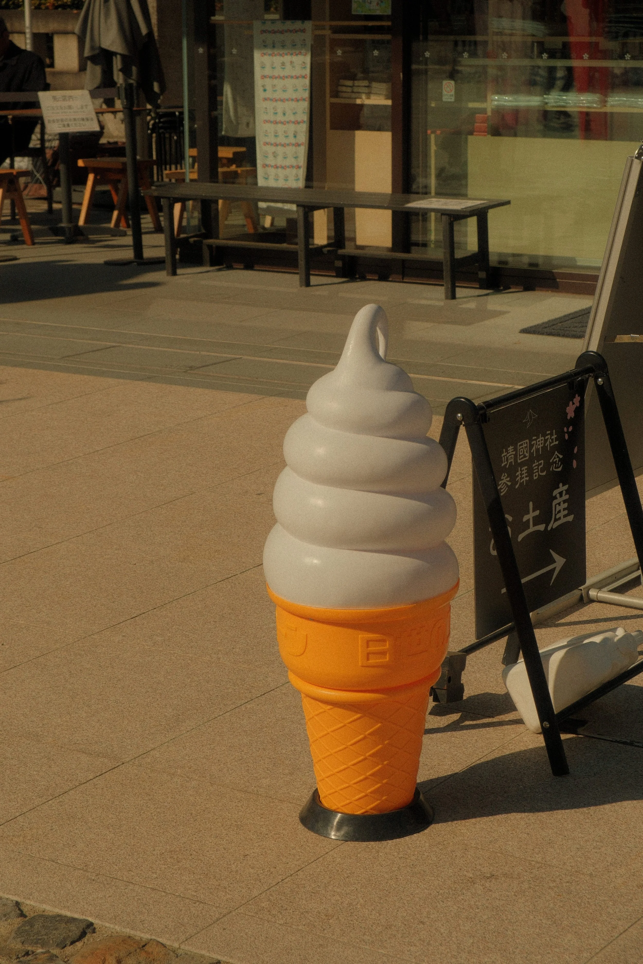 Large sculpture of a vanilla soft serve ice cream cone with an orange cone, placed outside on the sidewalk near a building with glass windows and a signboard.