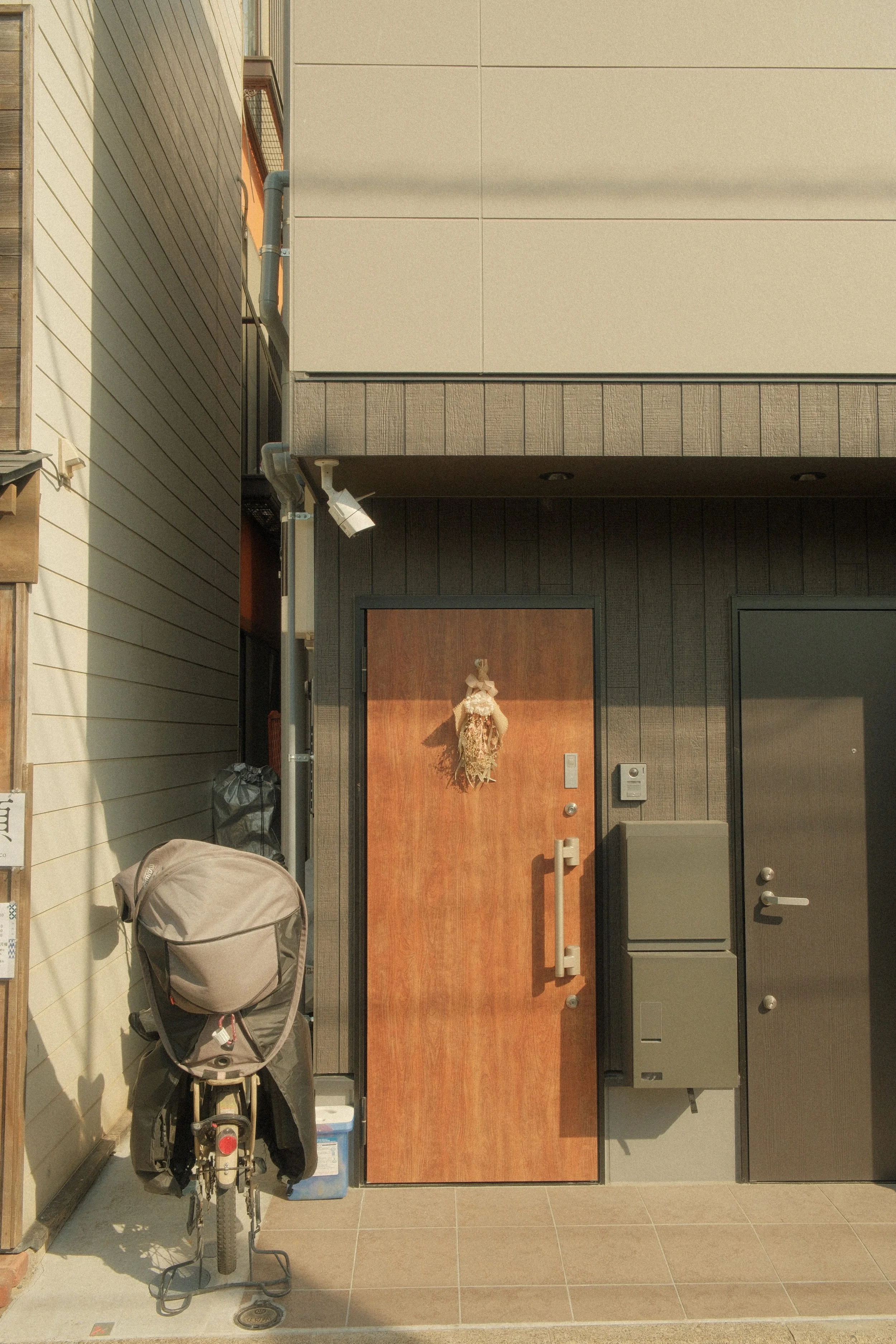 Exterior view of a house entrance with a wooden door decorated with a hanging Christmas wreath, a mailbox, intercom, and a closed door to the right. A bicycle with a large backpack is parked in front of the house.