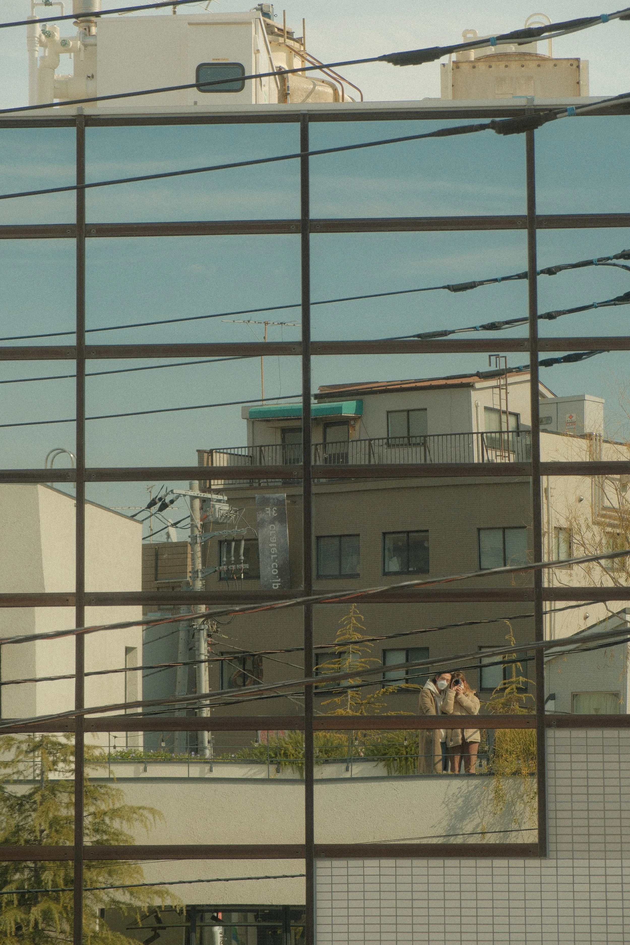A cityscape seen through a broken glass window, with reflections of two people taking a photo of the scene.
