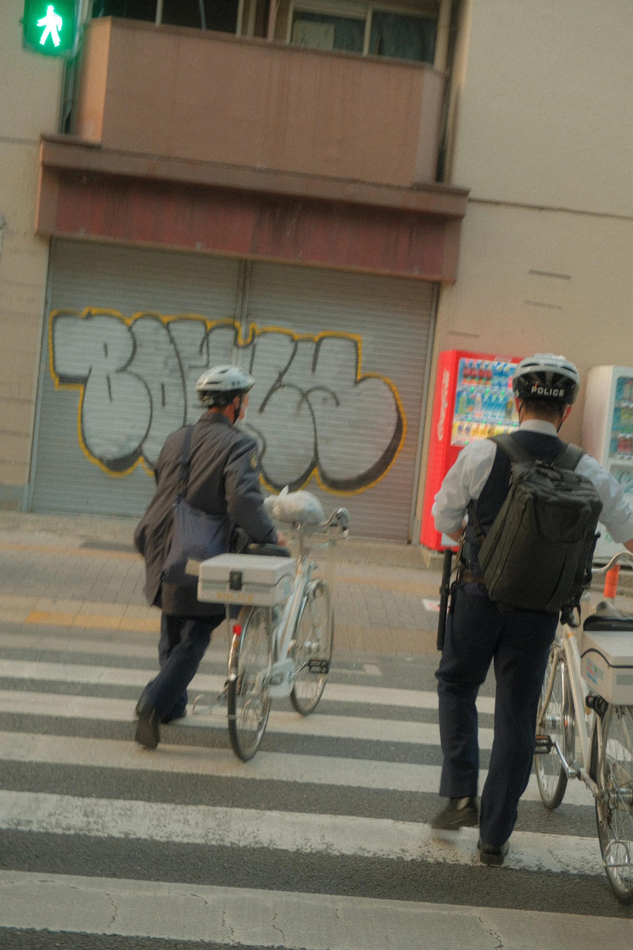 Two police officers on bicycles crossing a street with a graffiti-covered shutter in the background. One officer is wearing a backpack, and both are wearing helmets.