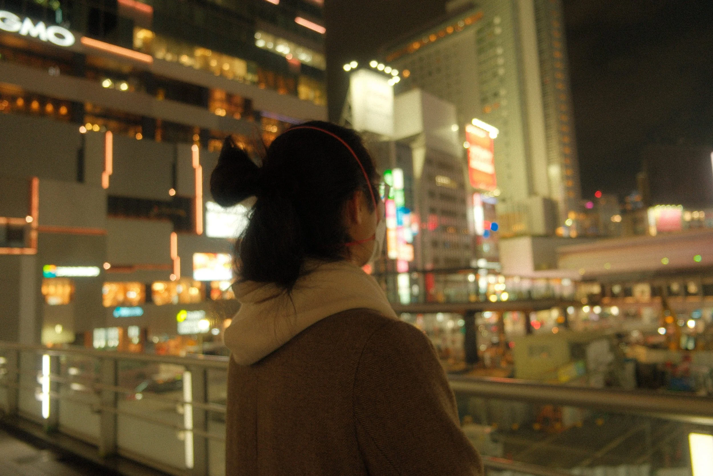 A woman with a bun hairstyle and wearing a mask looking at city lights at night from an elevated walkway.