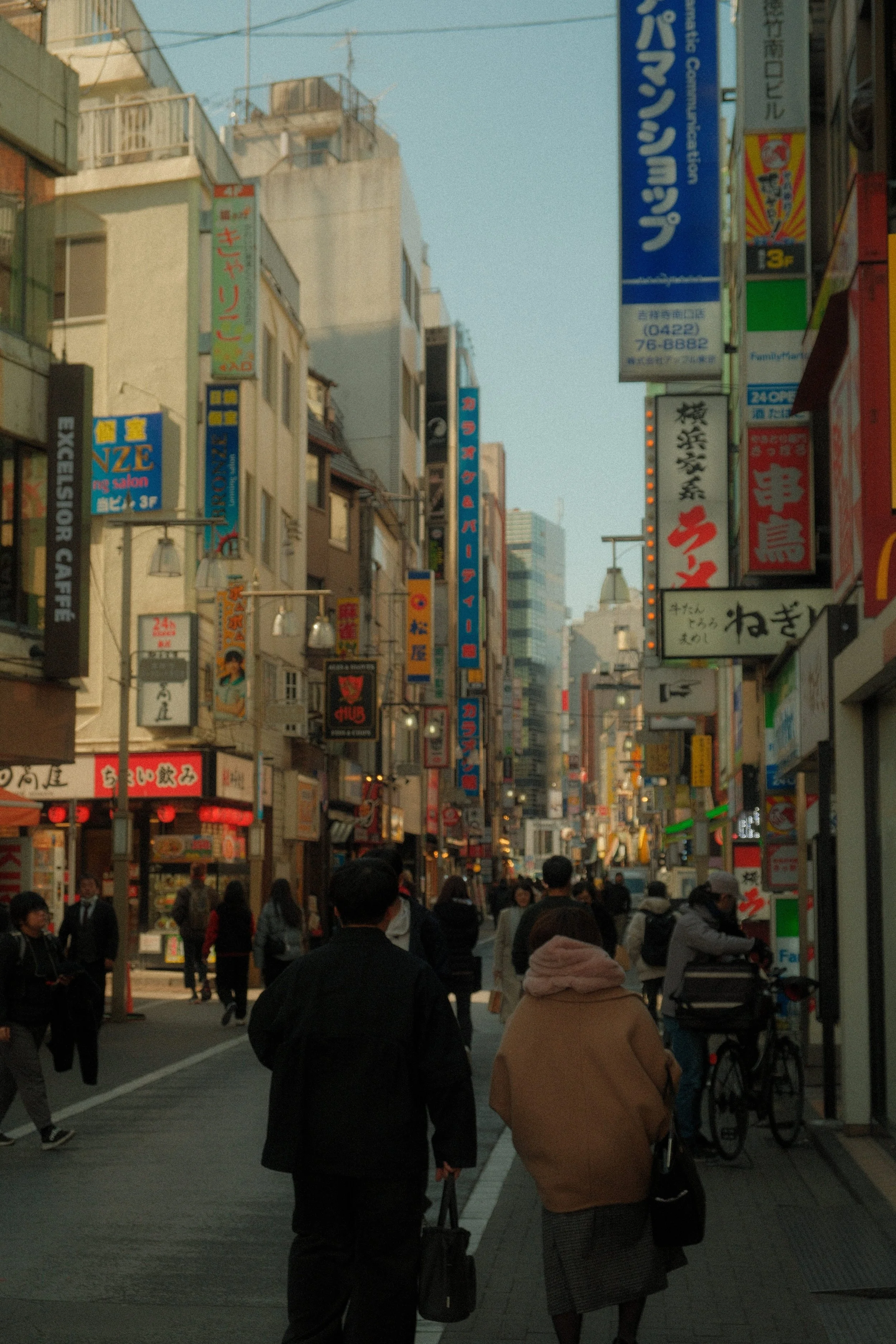 A busy urban street scene in Japan with people walking, and buildings with colorful signage and advertisements in Japanese.