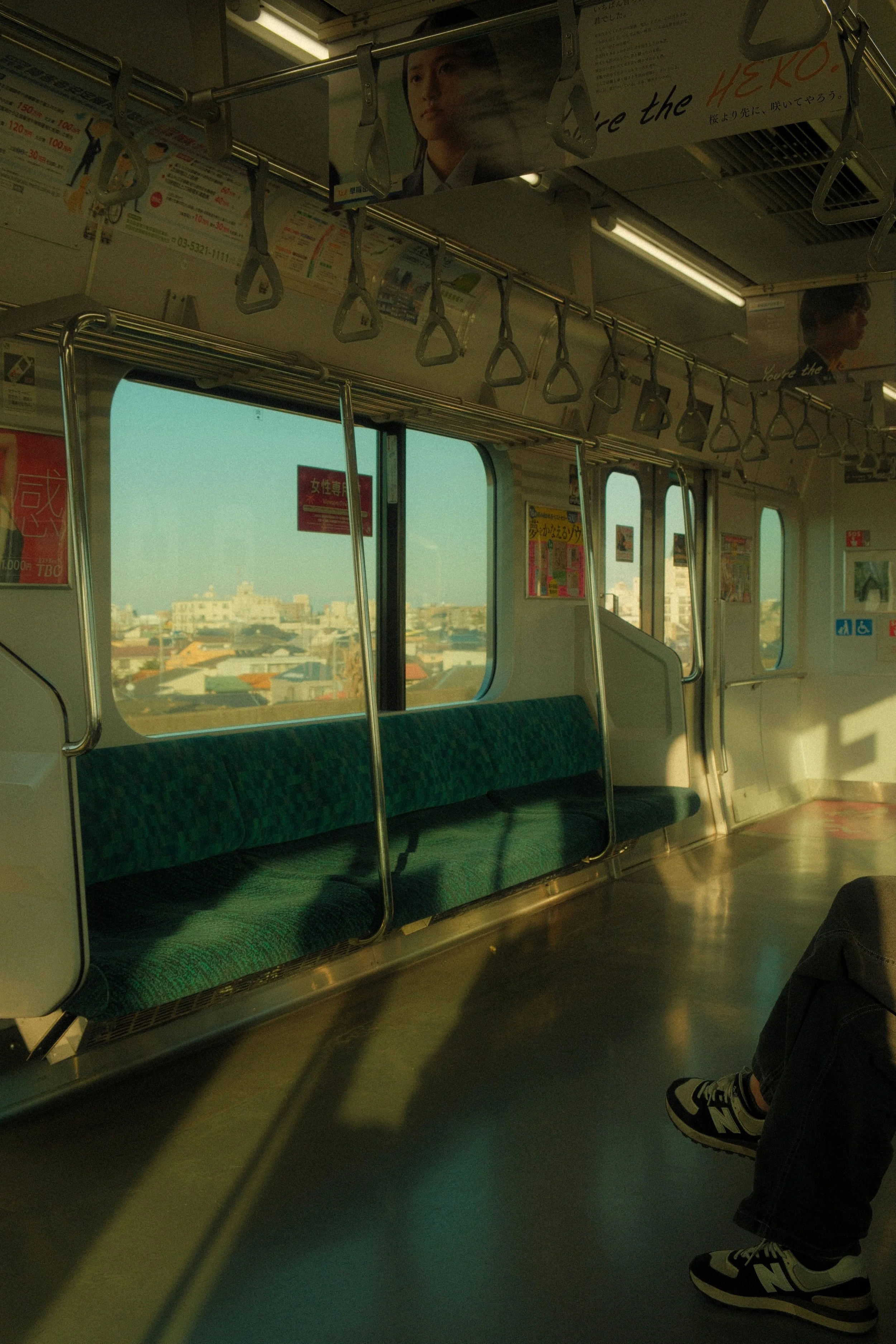 Empty subway train carriage with green patterned seats, large windows showing city buildings, and hanging hand straps for passengers to hold on to.