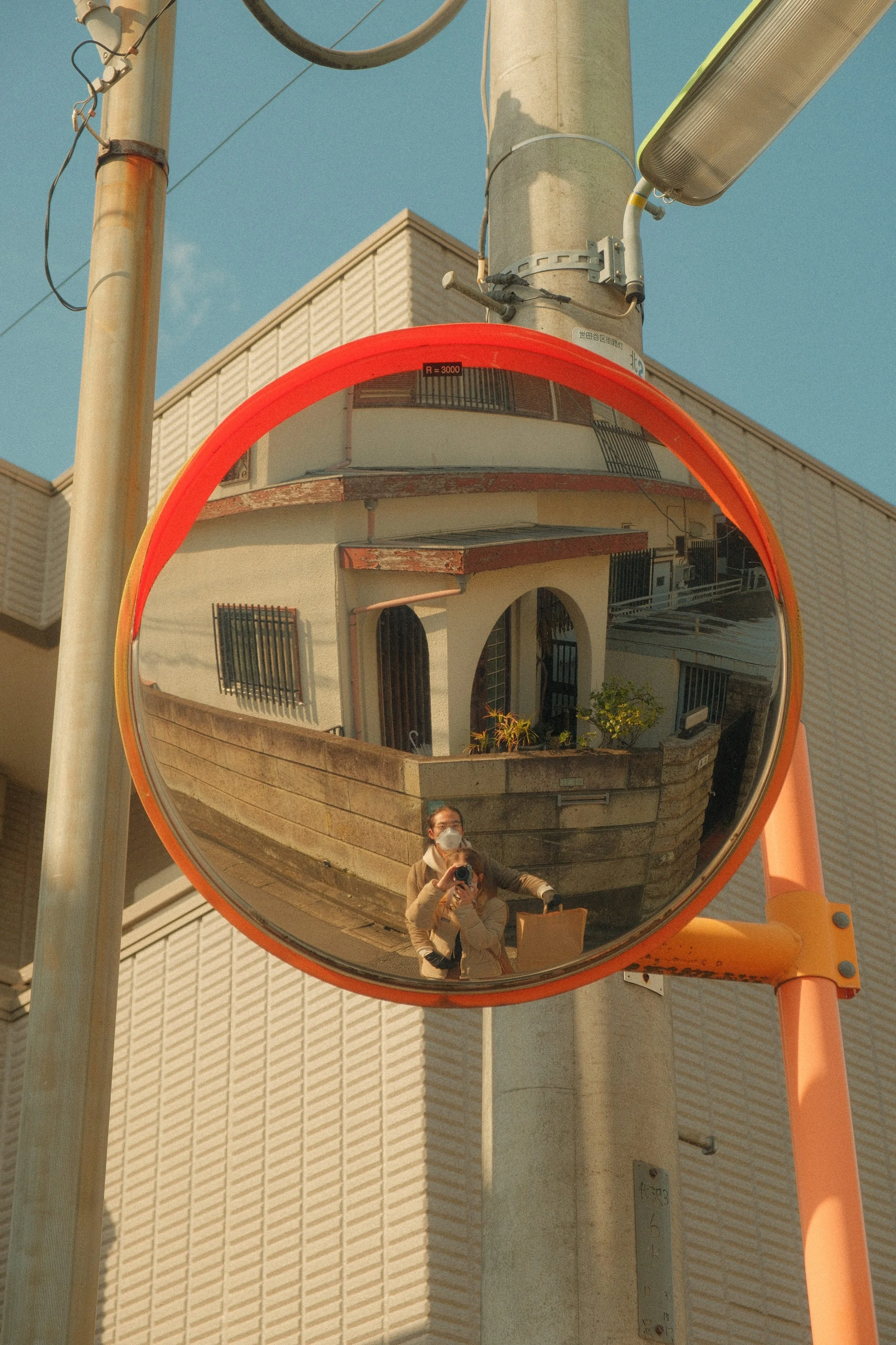 A convex traffic mirror reflecting a woman taking a photograph of herself, wearing a face mask, with buildings and a clear blue sky behind her.