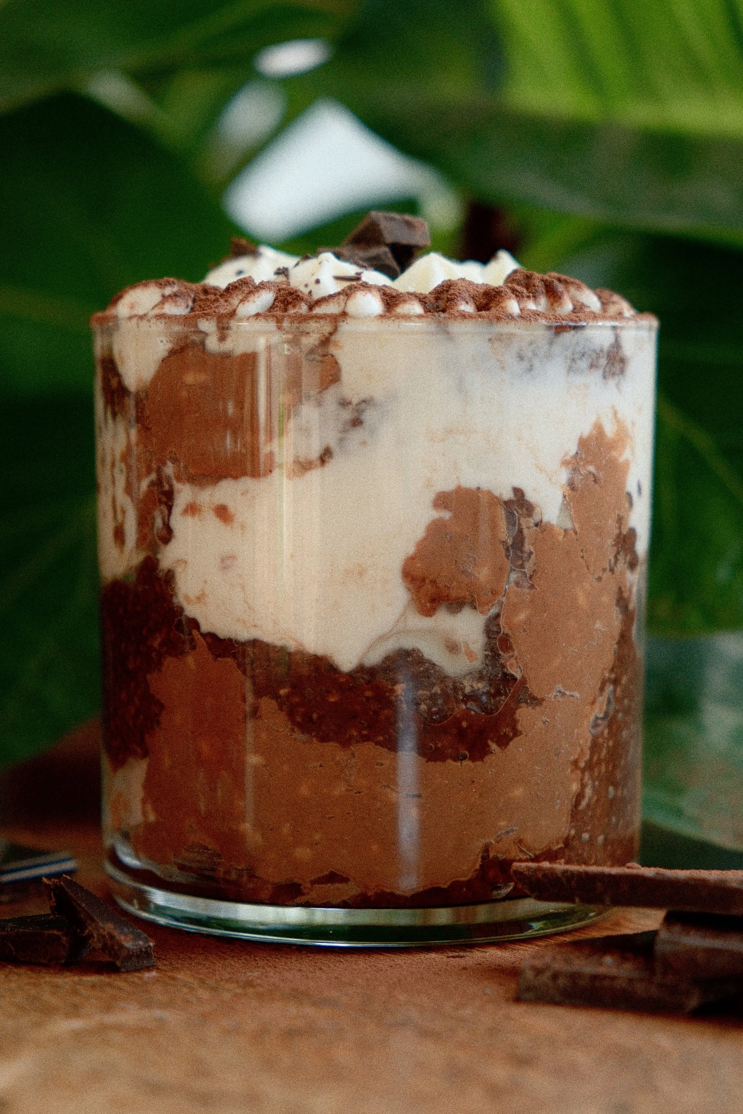 A layered chocolate and vanilla layered dessert in a glass with whipped cream and chocolate chunks on top, set on a wooden surface with dark chocolate pieces in the foreground and green leaves in the background.