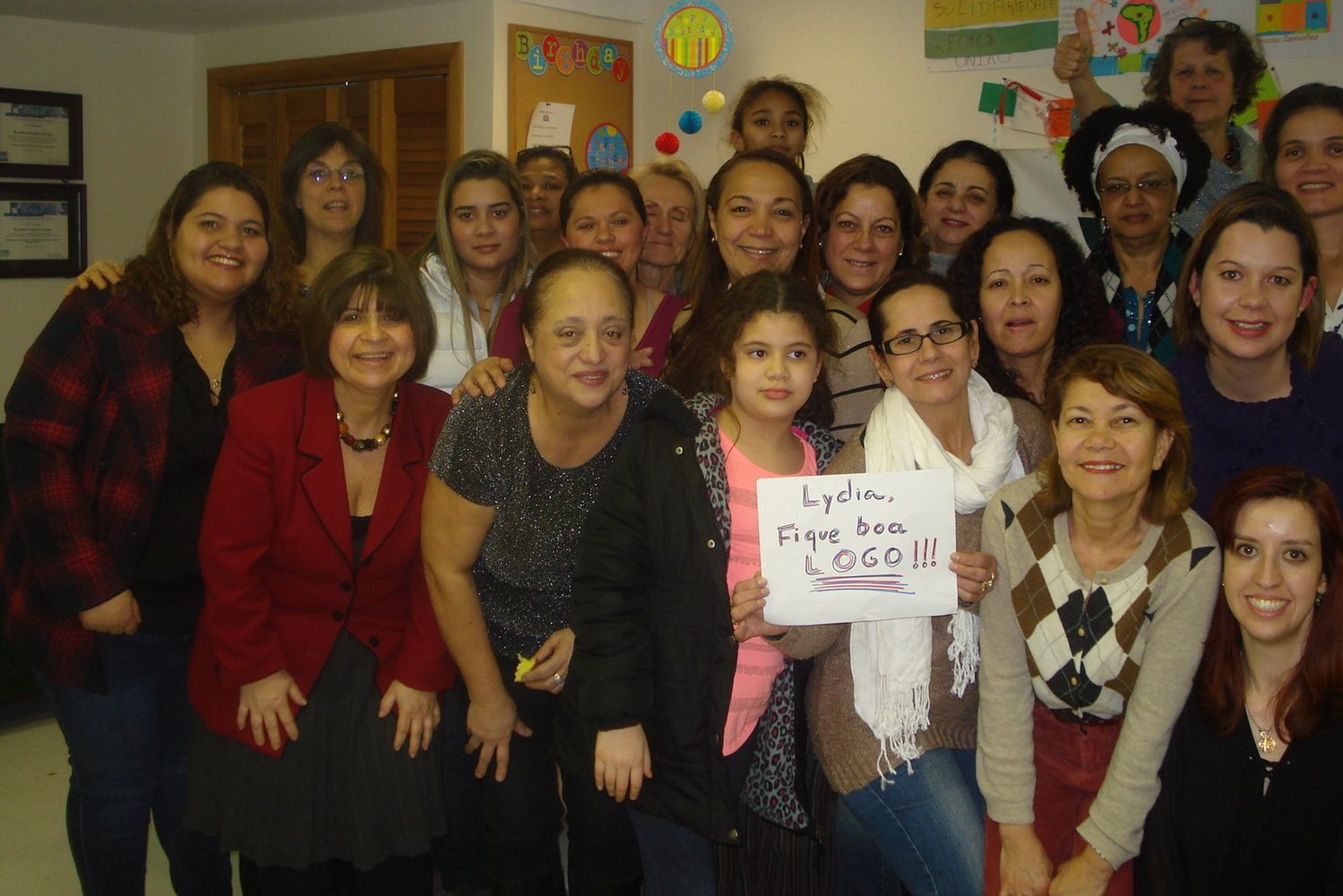  Más de veinte mujeres y niñas sonriendo a la cámara. Una al frente sostiene un letrero que dice "Lydia, Fique boa LOGO!!!" 