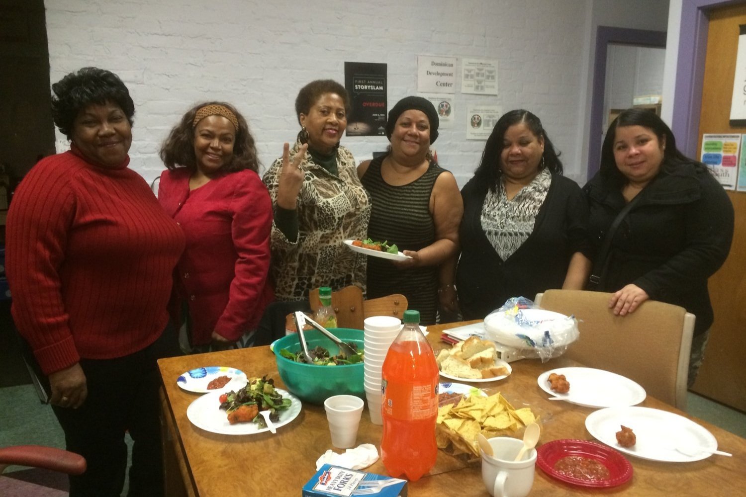  Seis mulheres de cor em volta de uma mesa, sorrindo para a câmera. Há comida na mesa. 