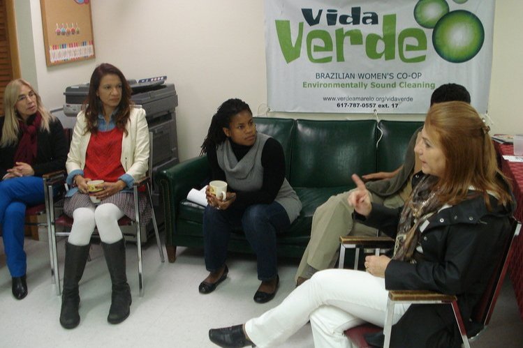  Cinco personas sentadas en círculo en sillas y un sofá verde. Un cartel en el fondo dice: “Vida Verde. Cooperativa de Mujeres Brasileñas. Limpieza ambientalmente saludable”. 