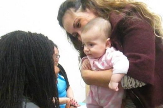  Mulher segurando um bebê em um macacão rosa. O bebê está olhando para o rosto de outra mulher, e as três parecem felizes. 