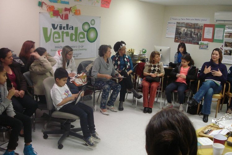  Muitas pessoas, incluindo várias crianças, sentadas em círculo em cadeiras e um sofá verde. Um pôster ao fundo diz: “Vida Verde. Cooperativa Feminina Brasileira. Limpeza Ambientalmente Saudável.” 