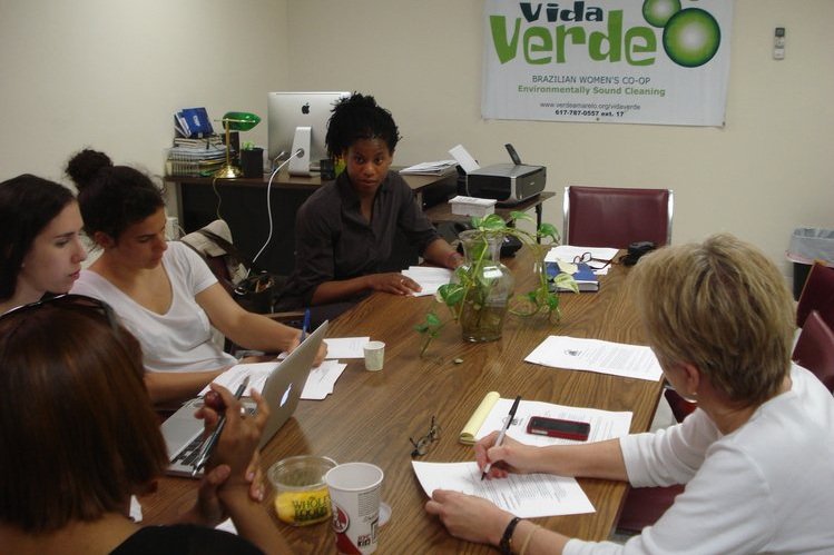  Cinco mulheres sentadas ao redor de uma mesa. Um pôster ao fundo diz: “Vida Verde. Cooperativa Feminina Brasileira. Limpeza Ambientalmente Saudável.” 