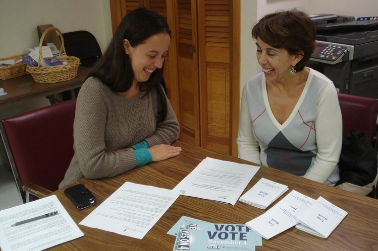  Two women sitting at a table facing each other and laughing. In the forefront of the image on the table, light blue cards are visible reading “Vote November 4. Yes on 4.” 