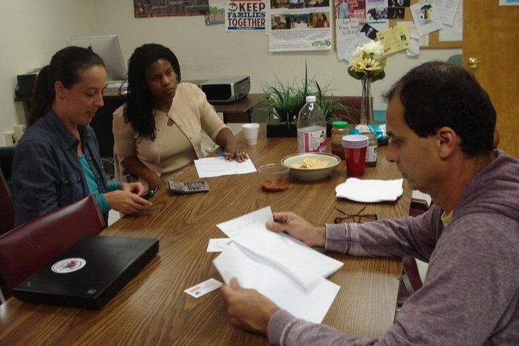  Three people sitting at a table in conversation. The man in the foreground is flipping through papers. 