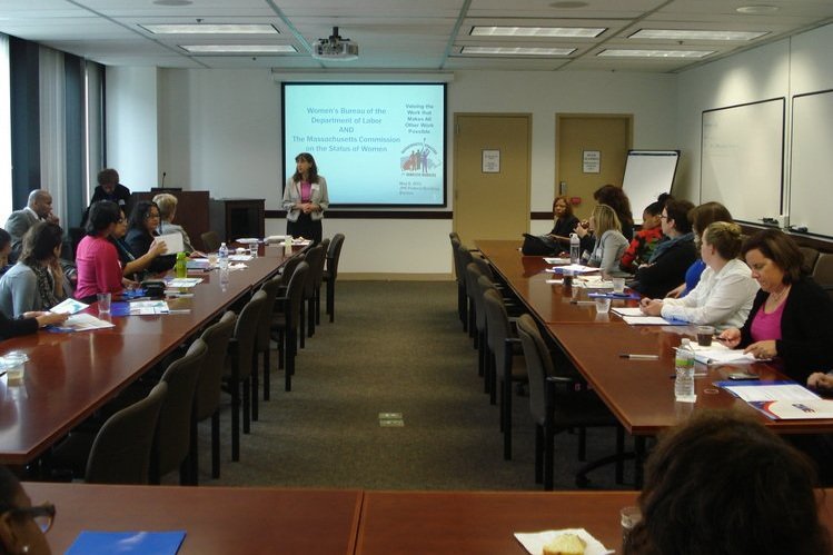  People at tables facing a presentation that reads, “Women’s Bureau of the Department of Labor and The Massachusetts Commission on the Status of Women. Valuing the work that makes all other work possible.” 