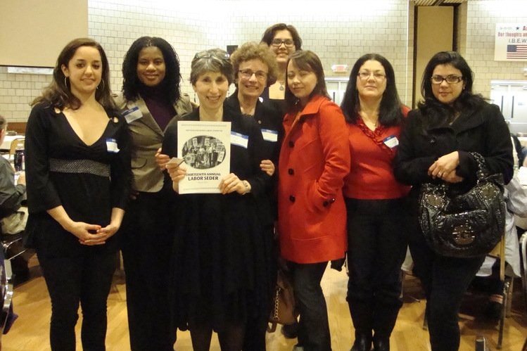  Oito mulheres juntas de frente para a câmera e sorrindo. Uma mulher na frente segura um papel onde se lê “Décimo Terceiro Seder Anual do Trabalho”. 