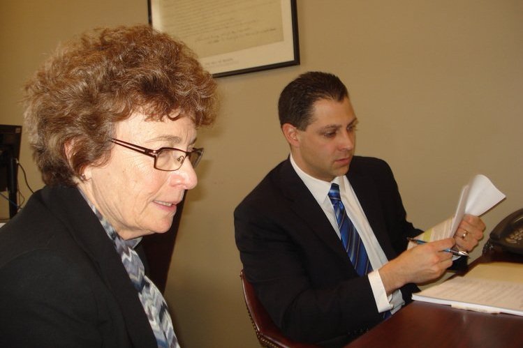  A woman and a man dressed in business attire sit at a desk. The man is reading through papers and is holding a pen. 