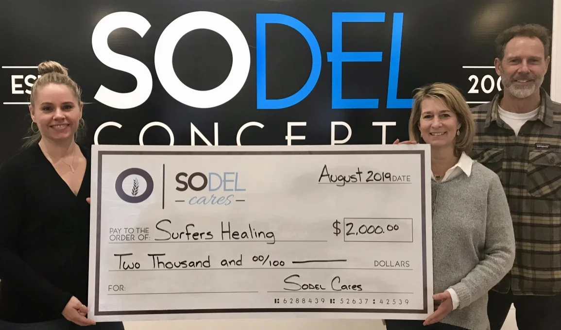 SoDel Cares, the philanthropic arm of SoDel Concepts, recently donated $2,000 to Surfers Healing, which uses surfing to enrich the lives of people with autism. Lindsey Barry, controller for SoDel Concepts, presents the check to Kelly and Dale Loeser, the owners of Quiet Storm, who help coordinate local Surfers Healing activities.