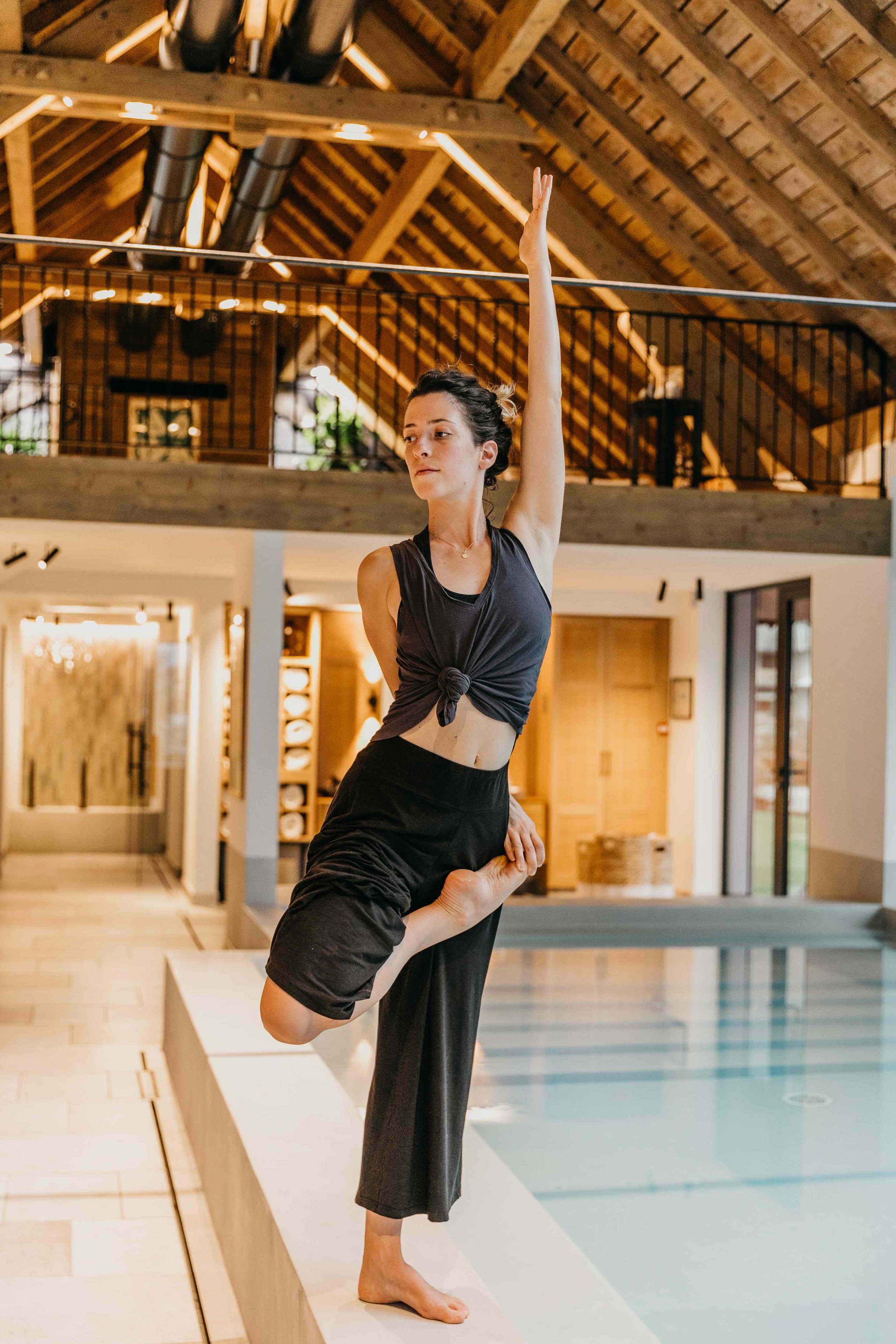 Une femme effectuant une posture de yoga ou de danse dans un intérieur avec plafond en bois et piscine.