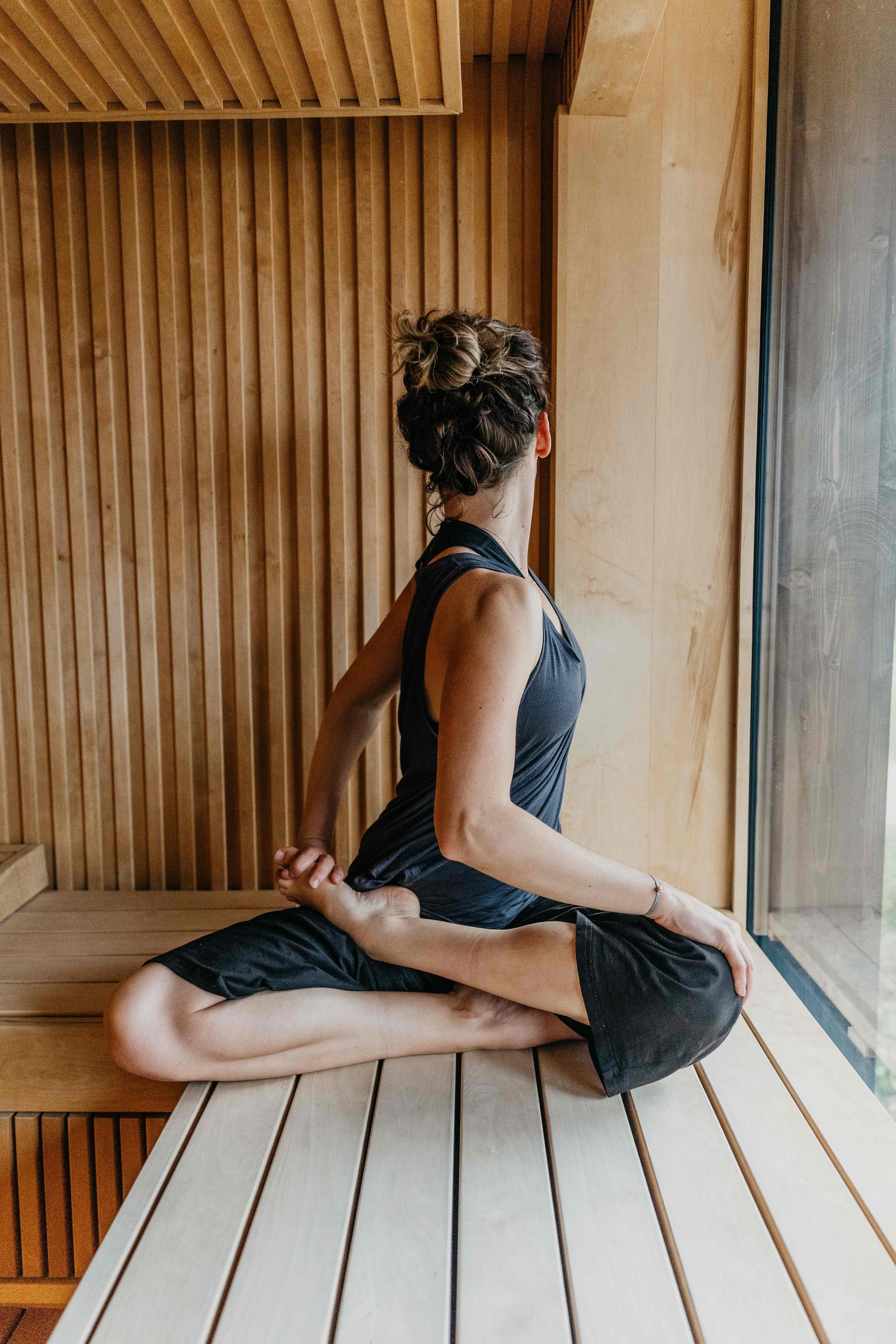 Femme pratiquant le yoga dans une pièce en bois, assise en position du héros offerte, regardant par une grande fenêtre.