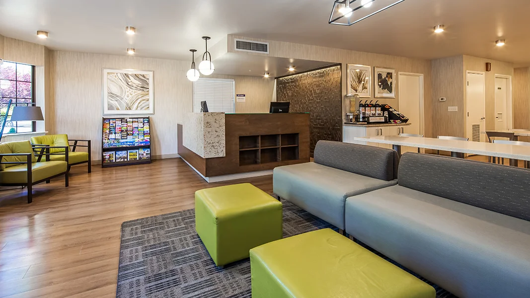 hotel lobby with gray and green soft seating, white breakfast area furniture, brown/ granite reception desk with accent wall