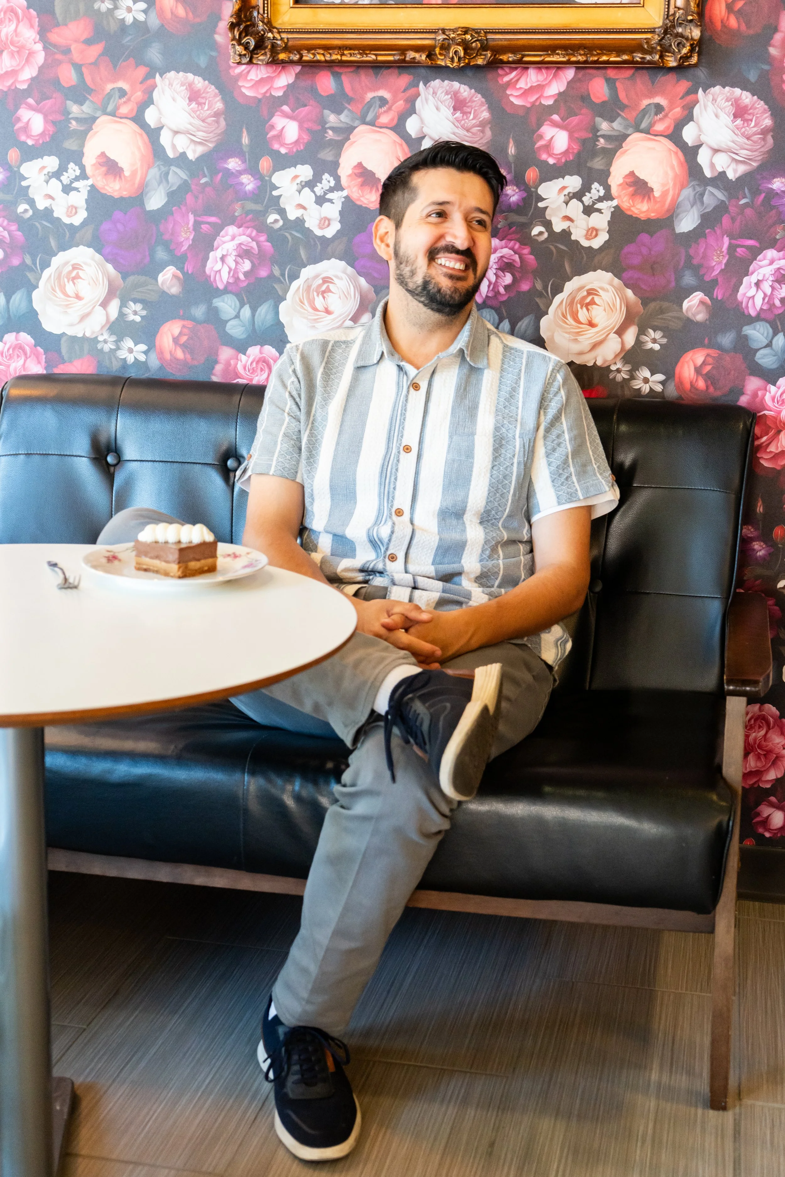 A man with dark hair and a beard sitting at a table in a restaurant with floral wallpaper. He is smiling, wearing a striped short-sleeve shirt, gray pants, and black sneakers. There is a piece of cake on a plate in front of him.