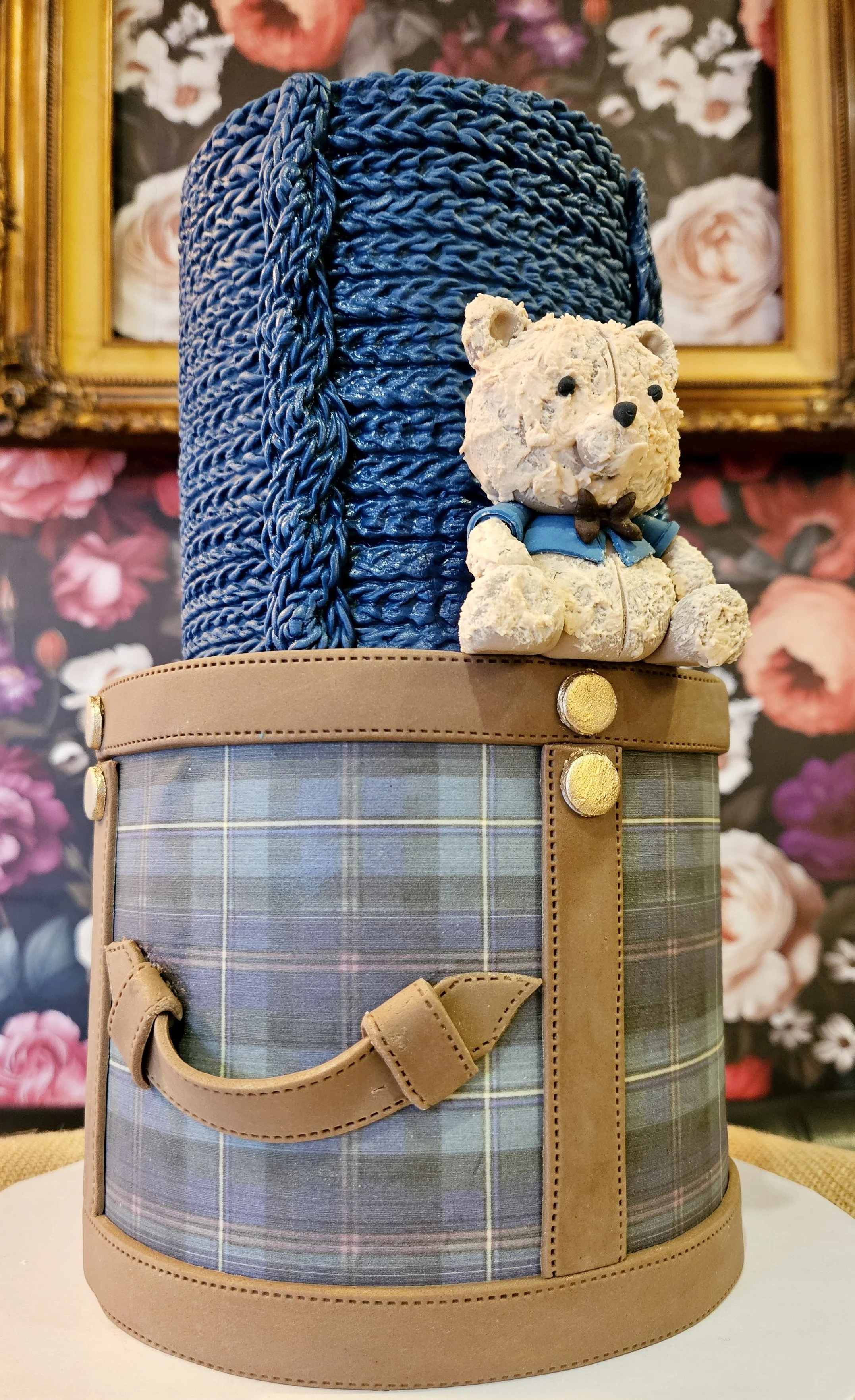 A teddy bear figure with a textured cream-colored body and a blue shirt, sitting in front of a large blue knitted hat. The scene features a patterned box with a plaid design and tan faux leather accents, set against a floral wallpaper background with a gold framed painting.