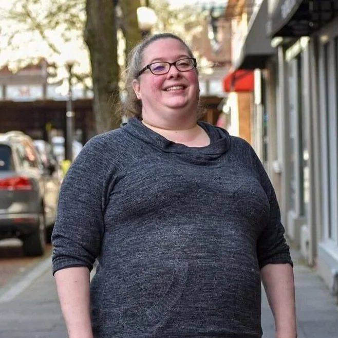 Person wearing glasses and a dark gray sweater, standing on a sidewalk with buildings and parked cars in the background.