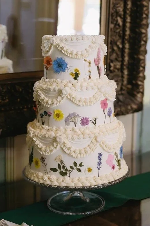 A three-tiered white wedding cake decorated with colorful flowers and white piped frosting swags, displayed on a glass stand.