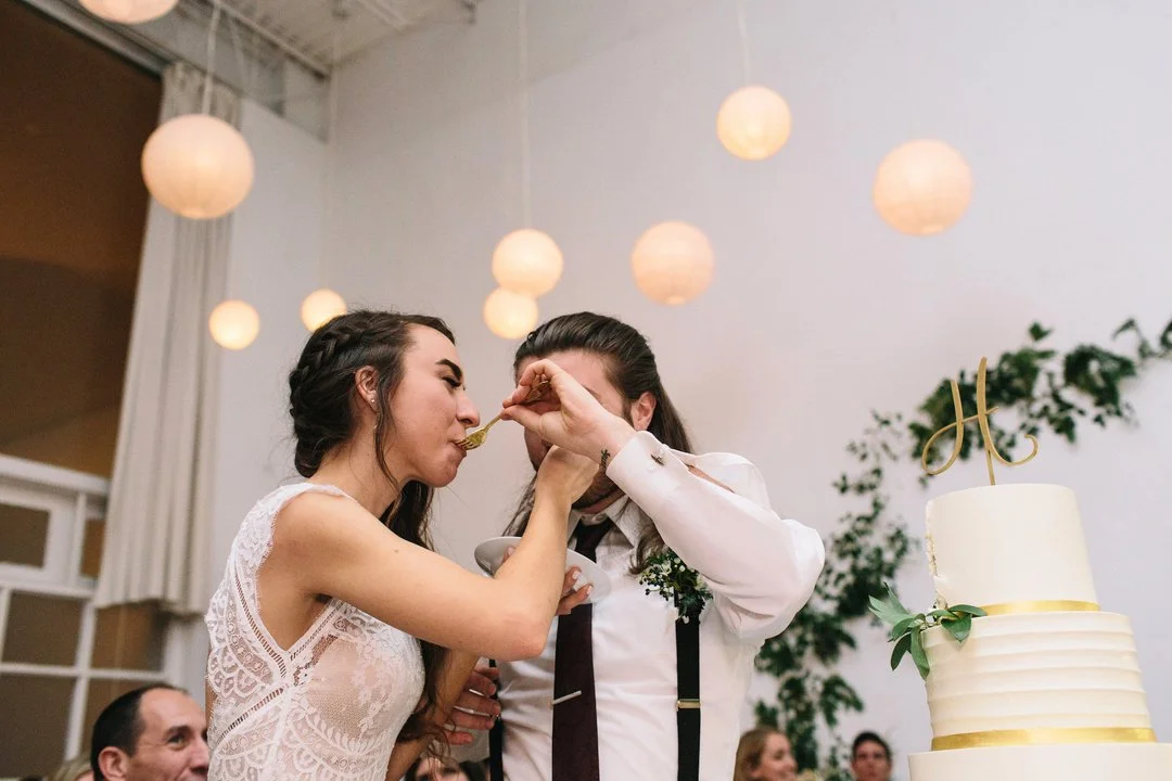 A bride and groom feed each other cake at their wedding reception. The bride is wearing a white lace dress, and the groom is in a white shirt with suspenders and a tie. There are paper lanterns hanging from the ceiling and a decorated wedding cake with a gold topper in the foreground.