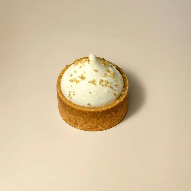 A small round tart with a golden brown crust filled with white cream and sprinkled with tiny gold flakes or crumbs, placed on a plain beige background.