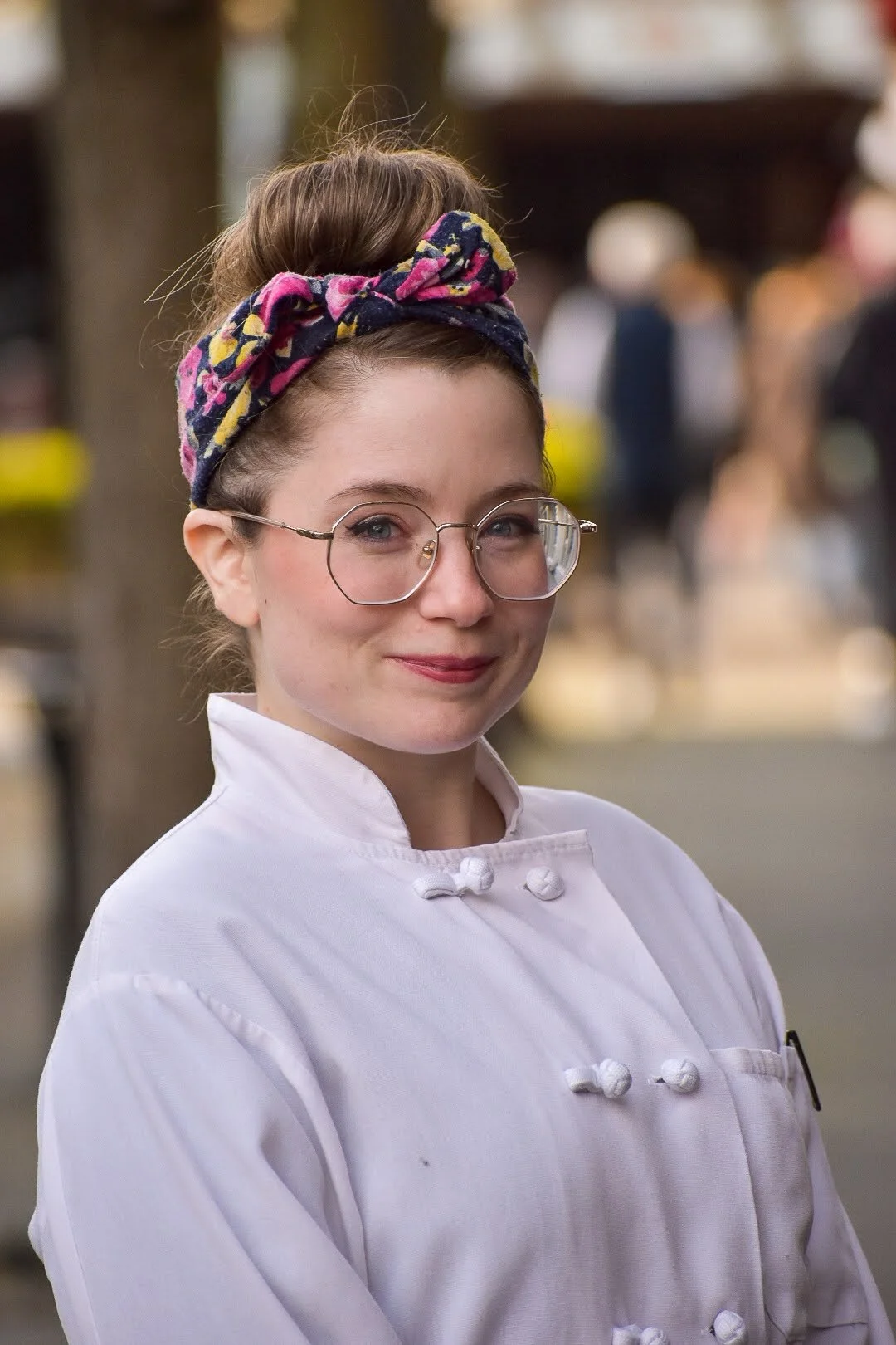 A woman wearing a white chef's coat, glasses, and a colorful headband, smiling at the camera.