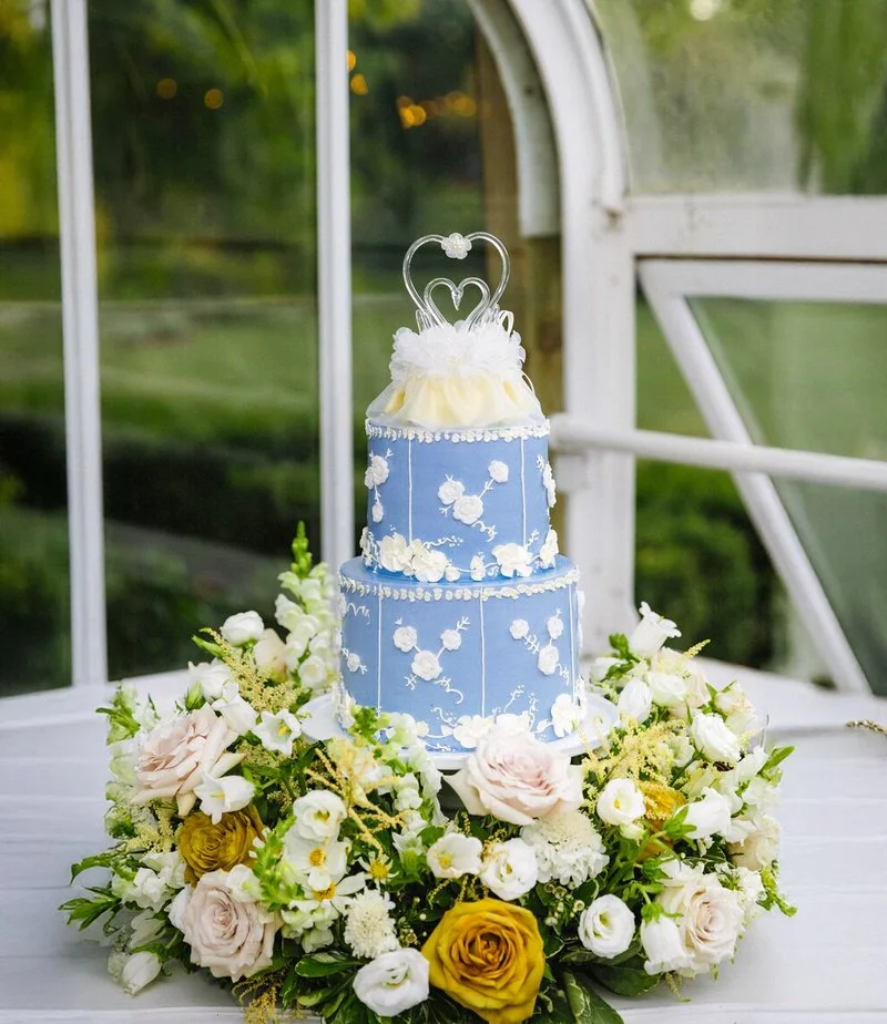 A three-tiered blue wedding cake decorated with white floral patterns, topped with a heart-shaped topper, sits on a white table surrounded by a bouquet of white, cream, and yellow flowers.