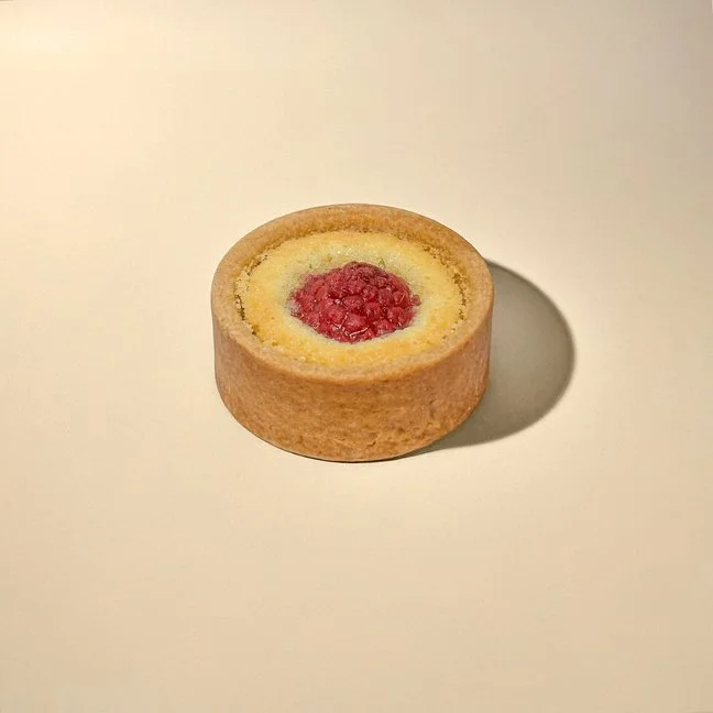 A round cookie with a yellow cake-like center and a raspberry in the middle, placed on a plain white surface, casting a shadow.