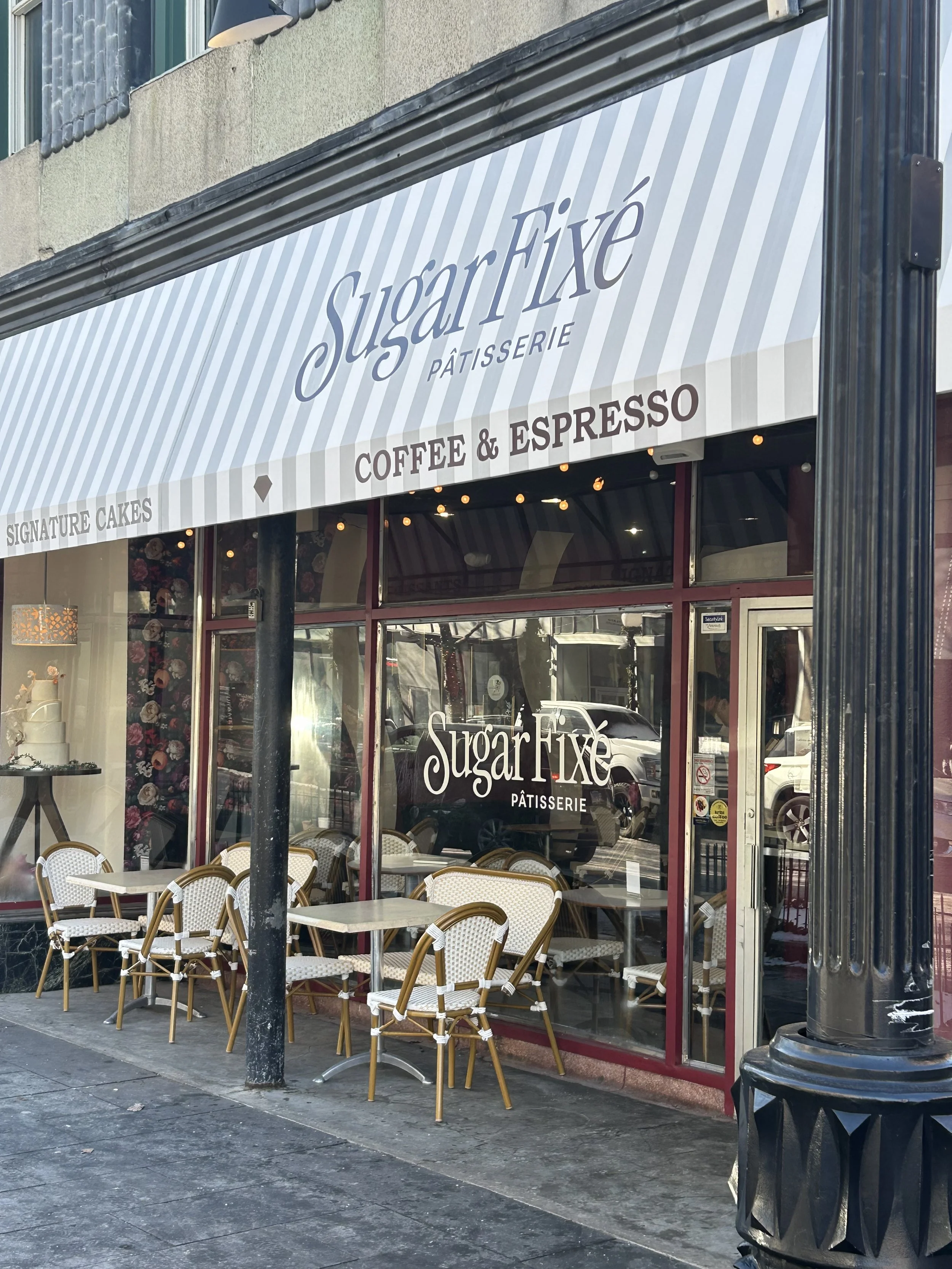 Exterior of a bakery or café named Sugar Fixe Pâtisserie with outdoor seating, glass windows, and an awning. The sidewalk is visible with chairs and tables outside.