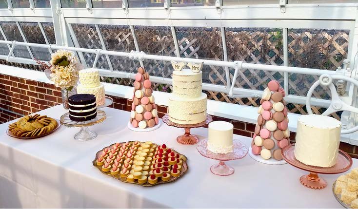 A dessert table with various cakes and treats, including macarons, cookies, and layered cakes, set up outdoors on a white table with a white railing and building in the background.