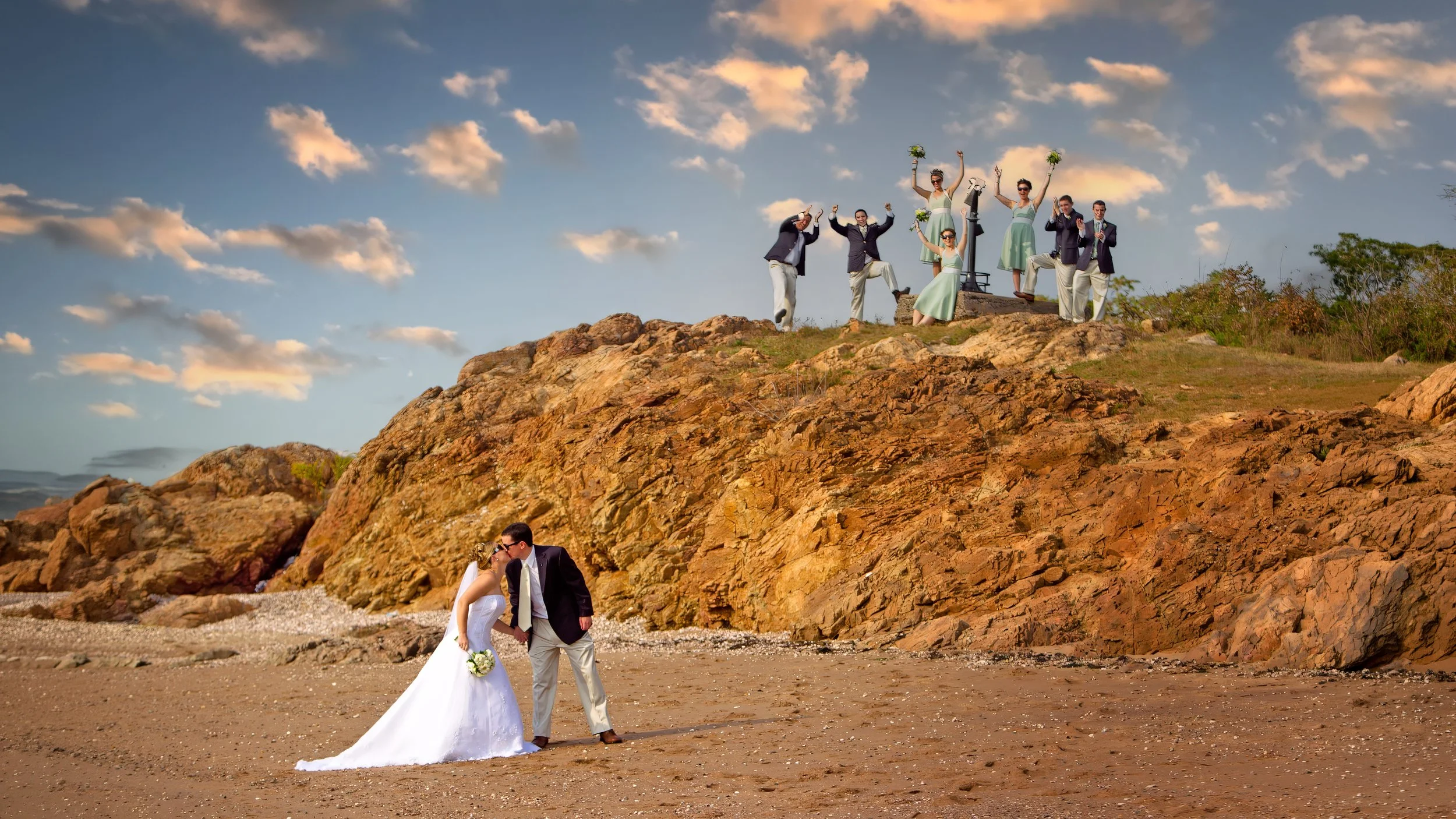 Bride and groom kissing on the beach at Lighthouse Point Park with wedding party cheering from the cliffs above in New Haven Connecticut