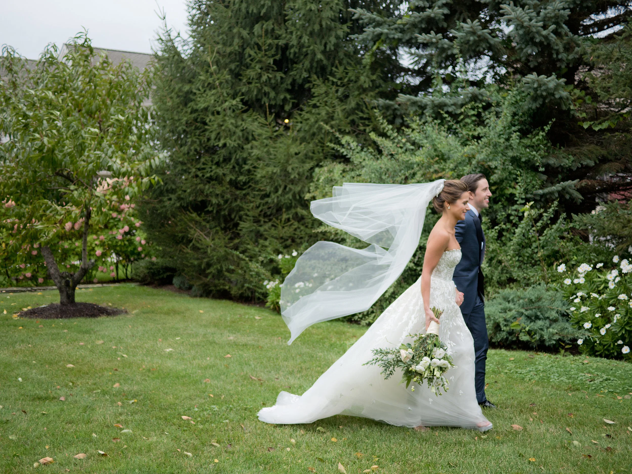 Bride and groom walking through garden lawn with veil blowing in wind at Lion Rock Farm