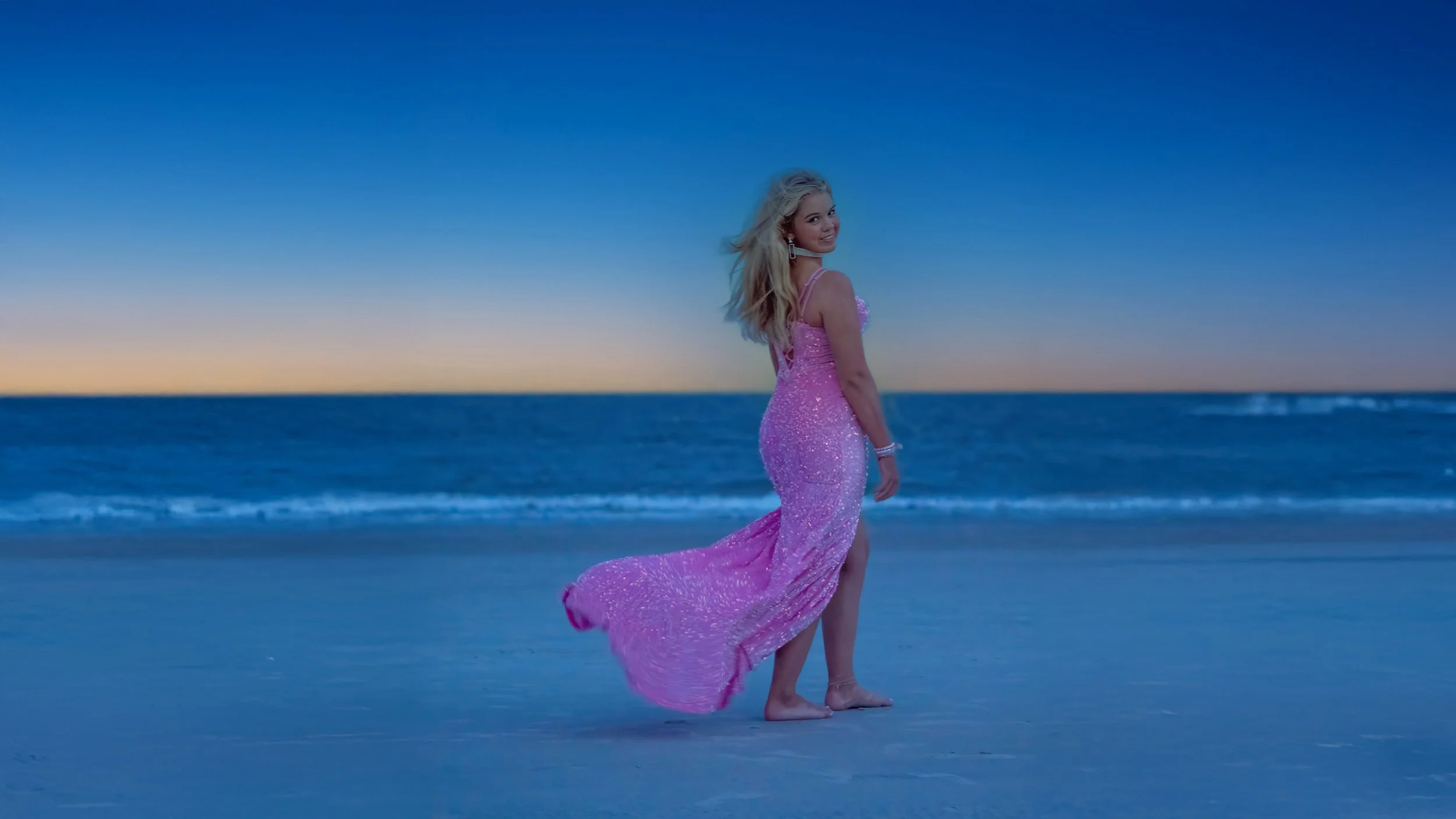 Senior portrait of a young woman in a flowing pink dress against a late sunset and twilight sky.
