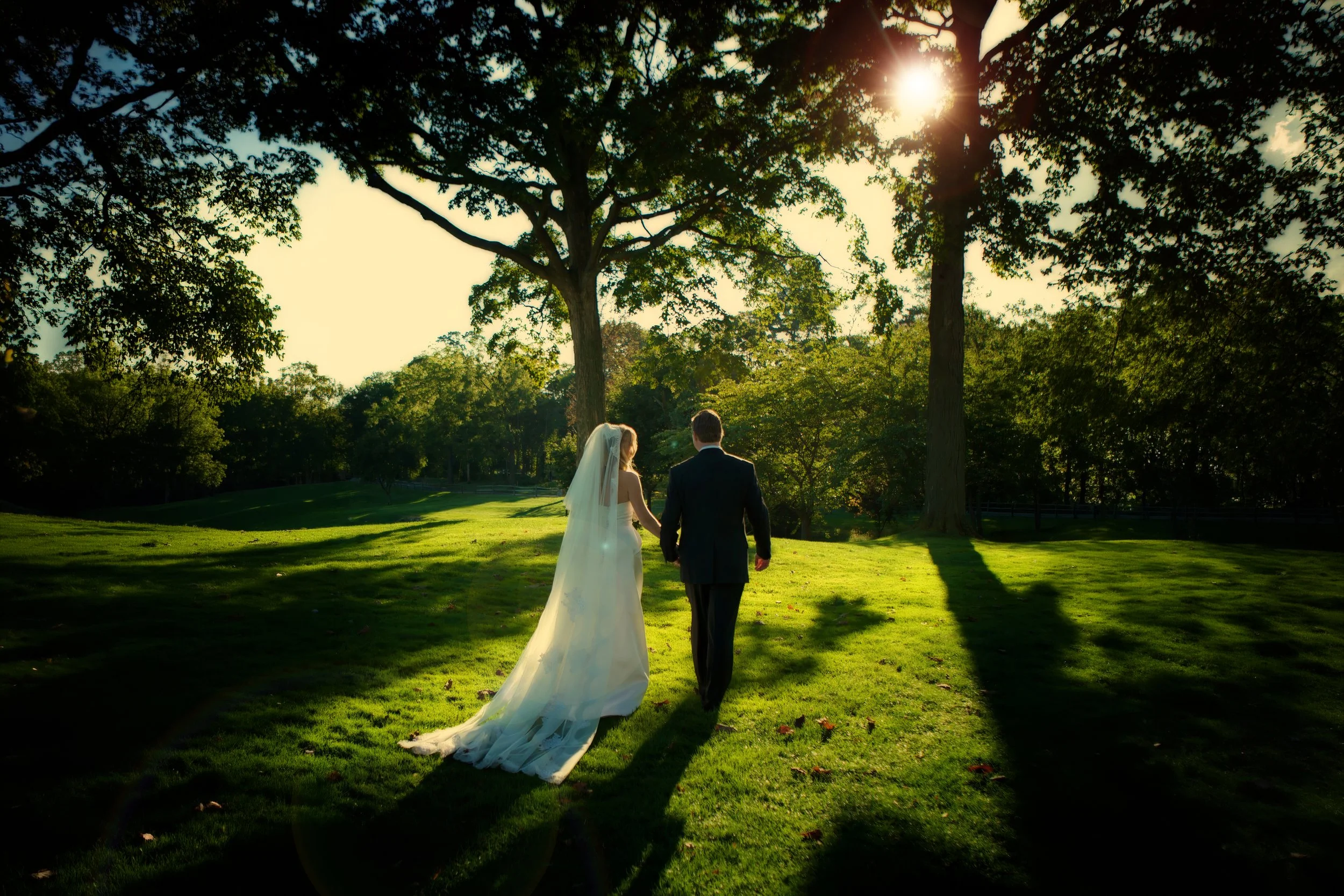 Bride and groom walk on the grounds of Siwanoy Country Club as golden sunlight filters through the trees.