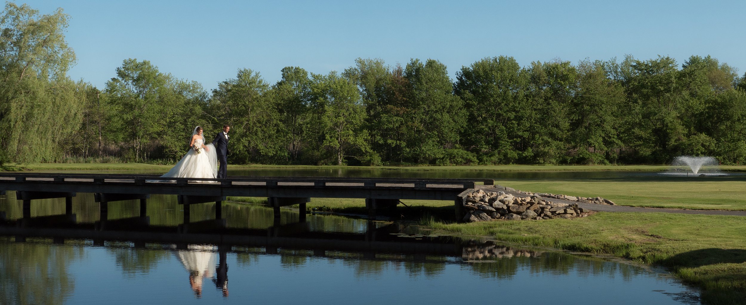 Bride and groom walk across a bridge at Salem Golf Club with their reflections mirrored in the water.