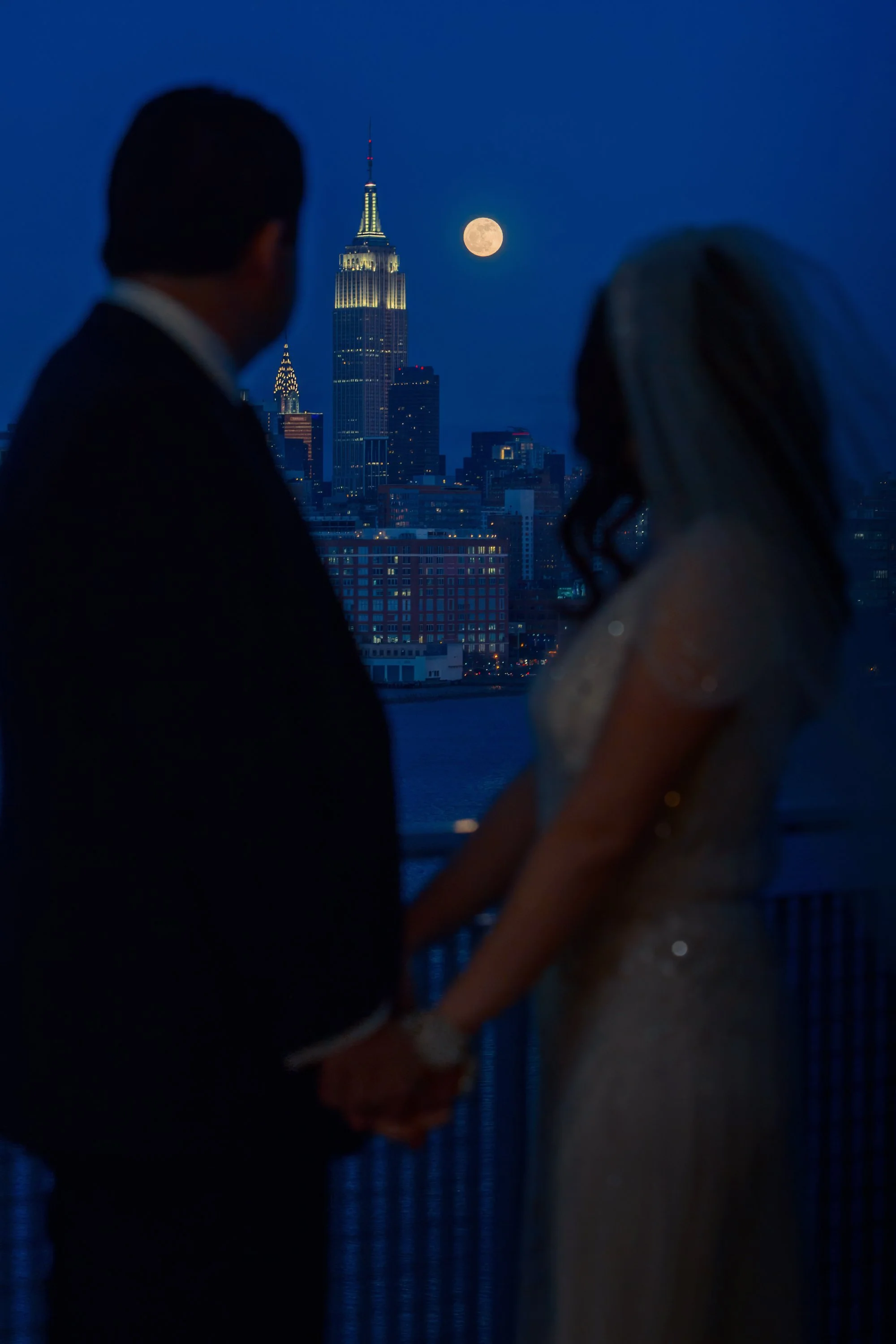 Bride and groom in silhouette gaze at the Manhattan skyline from W Hoboken at twilight with a full moon overhead.