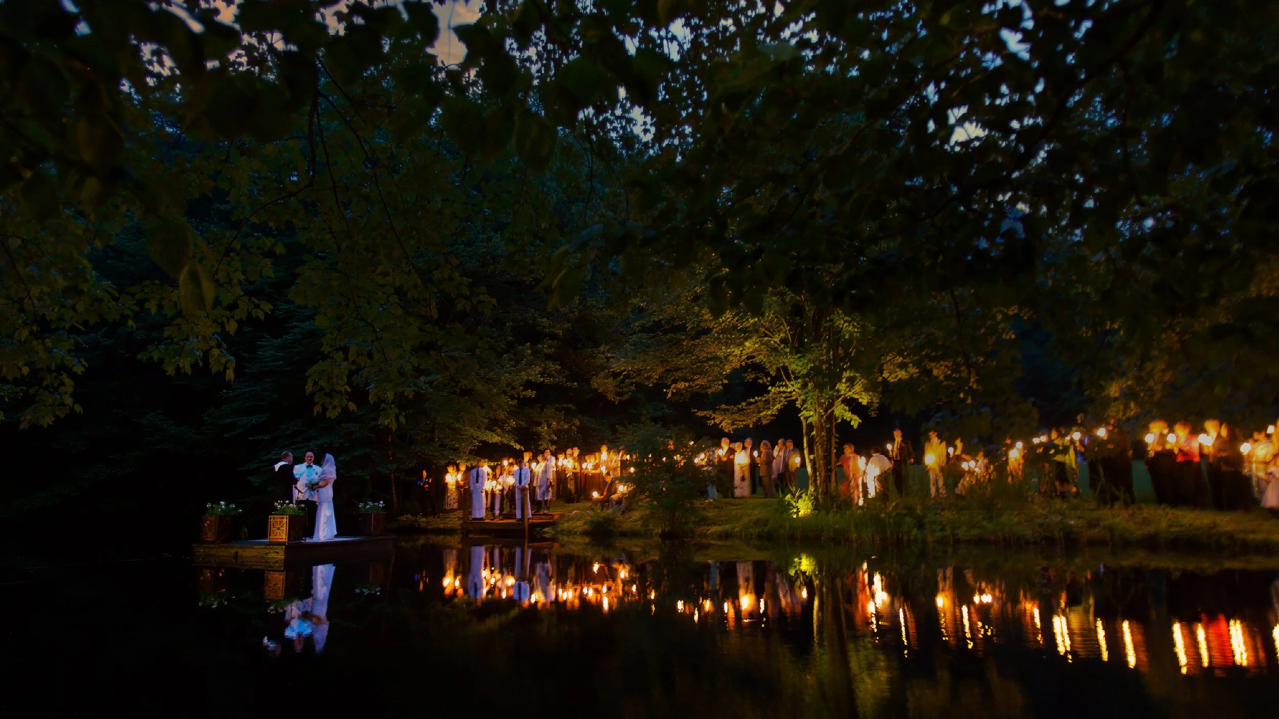 Wedding ceremony beside a pond in Connecticut at dusk with candlelight glowing along the water.